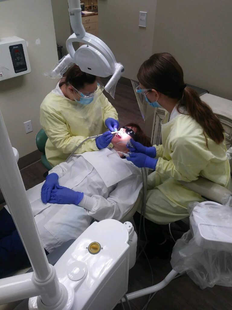Two women in yellow lab coats and blue gloves are working on a patient.