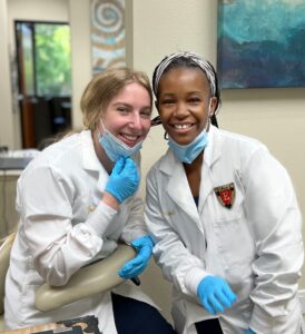 Two women in lab coats and blue gloves smile for the camera.