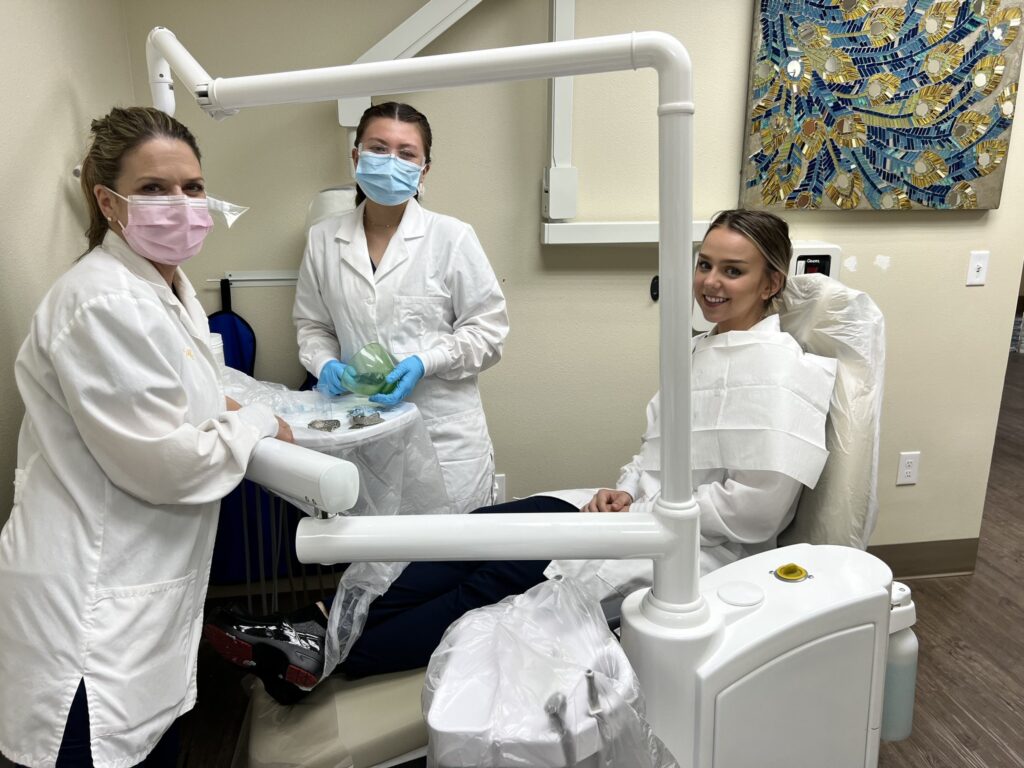 Three people in white lab coats and masks are standing near a dental chair.